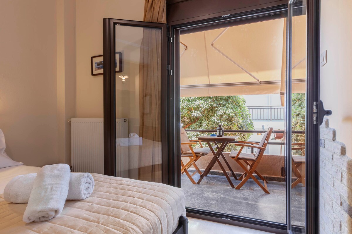 The interior features a spacious bed dressed with neutral linens. Glass doors open to a private balcony, with a small table and chairs visible outside. A sunshade provides shelter, while greenery in the background adds a natural touch.