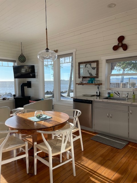 A cozy dining area features a round wooden table surrounded by four white chairs. Natural light fills the space through large windows, showcasing views of the serene waterfront. A compact kitchen is equipped with modern appliances, while a television is mounted on the wall for entertainment.
