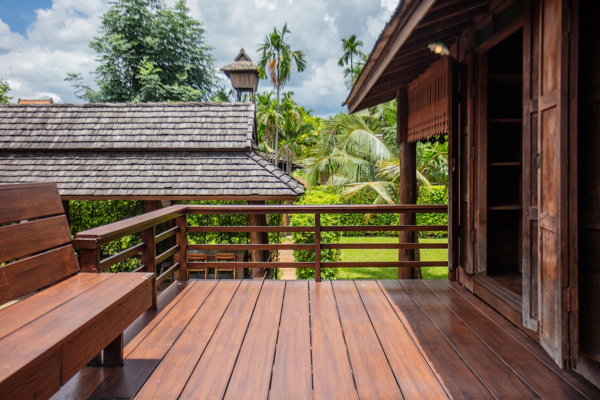 A wooden deck stretches out before the entrance, furnished with a simple bench. Lush greenery and palm trees are visible in the background, enhancing the serene environment. Soft clouds float overhead, creating a peaceful atmosphere.