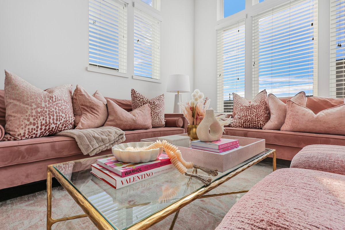 A bright living area is adorned with soft pink sofas featuring multiple decorative cushions. A glass coffee table displays art books and a neutral decorative piece. The space is illuminated by natural light streaming through large windows with white blinds.
