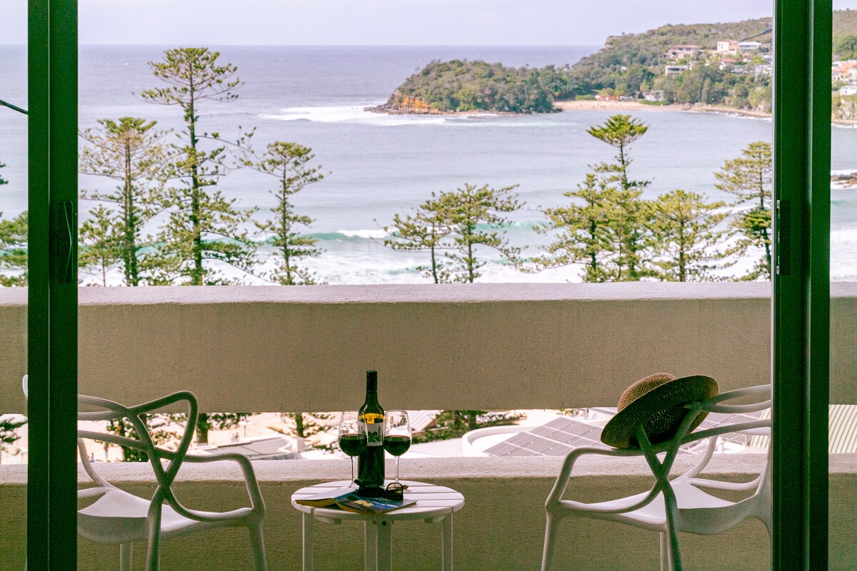 The balcony view showcases a stunning coastline with rolling waves and a lush hillside. Two white armchairs and a small table sit invitingly, complemented by a bottle of wine and glasses, surrounded by green trees that frame the picturesque setting.