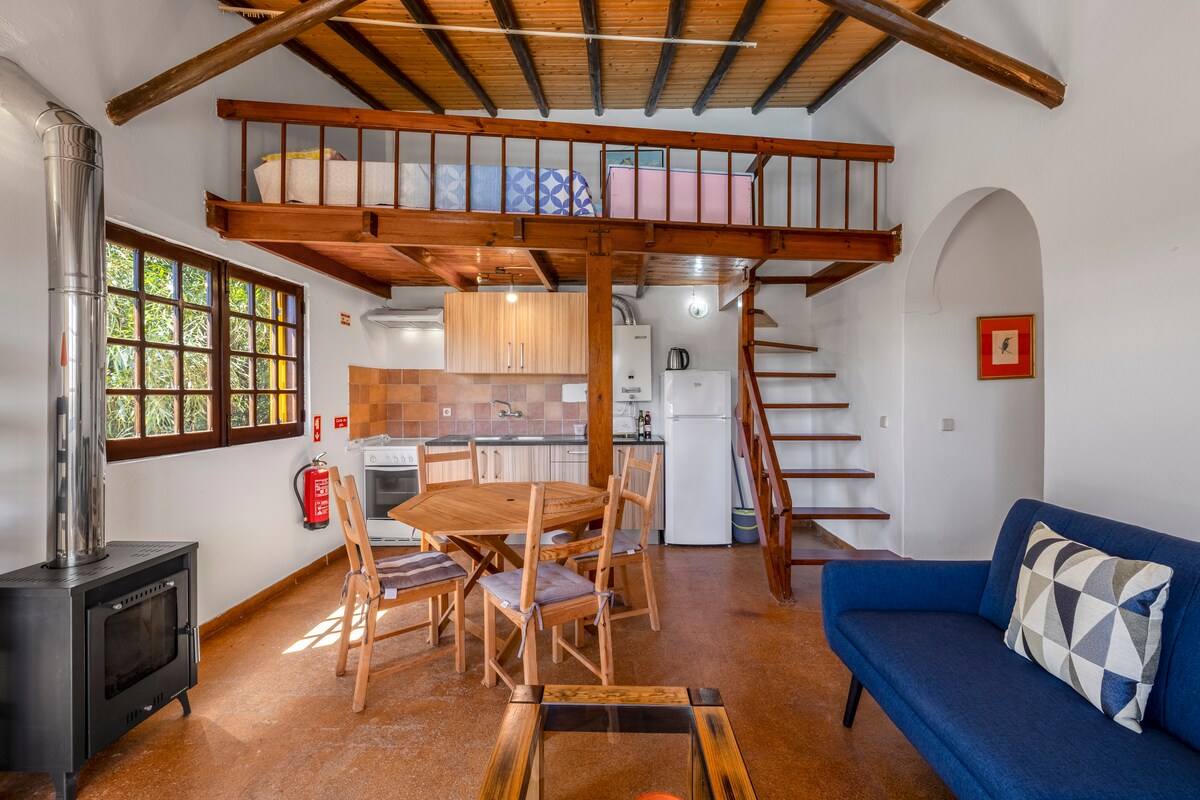 An open living space features a wooden ceiling with exposed beams. A round dining table accommodates several chairs. A blue sofa adds comfort, while a staircase leads to a loft area. A wood-burning stove is situated nearby, enhancing the room's warmth.