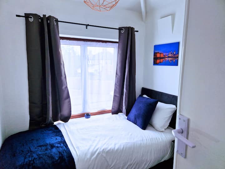 חדר שינה מזמין עם מיטה הממוקמת ליד חלון, עם וילונות בצבע חום, כריות לבנות וכחולות ותמונת קיר דקורטיבית, שיוצרים מקום מפלט שלו ואלגנטי