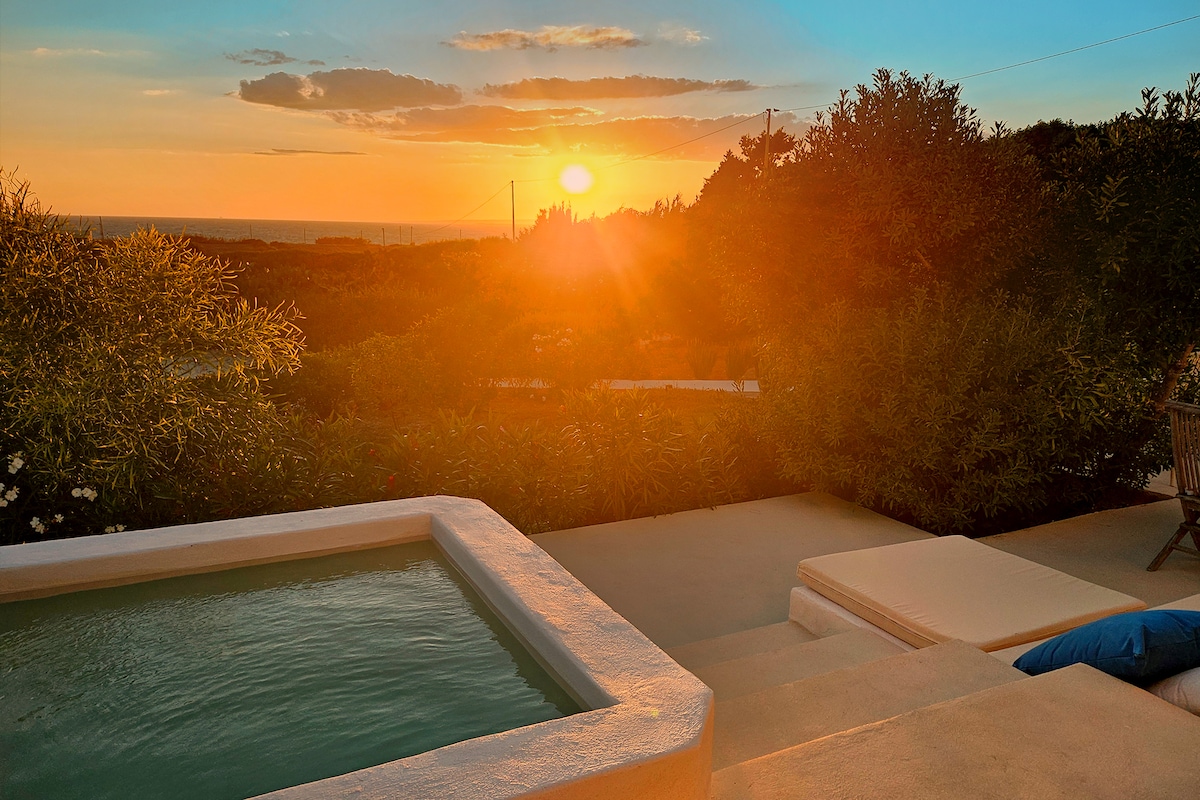 A panoramic view captures a vibrant sunset over the Mediterranean, framed by greenery. A private hot tub is visible alongside a lounge area with a light-coloured cushion, offering a peaceful space to enjoy the scenery.