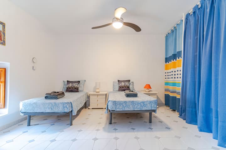 Spavaća soba s dva odvojena kreveta, stropnim ventilatorom, klima-uređajem i prozorom s drvenim griljama. Uključuje plave zavjese, stolić s lampama, toaletni stolić s ogledalom i stolicu. Prostran, udoban i dobro osvijetljen prostor.
