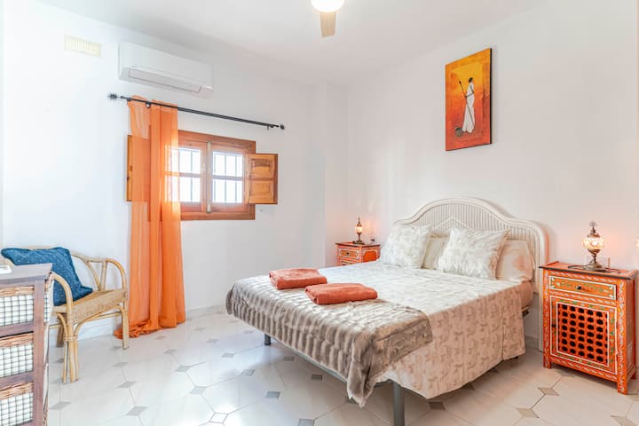 Spavaća soba s bračnim krevetom, ugrađenim ormarom, klima-uređajem i prozorom s narančastim zavjesama. Uključuje stolove s lampama, ogledalo, komodu i pletenu stolicu. Svijetao, funkcionalan i dobro opremljen prostor za odmor.
