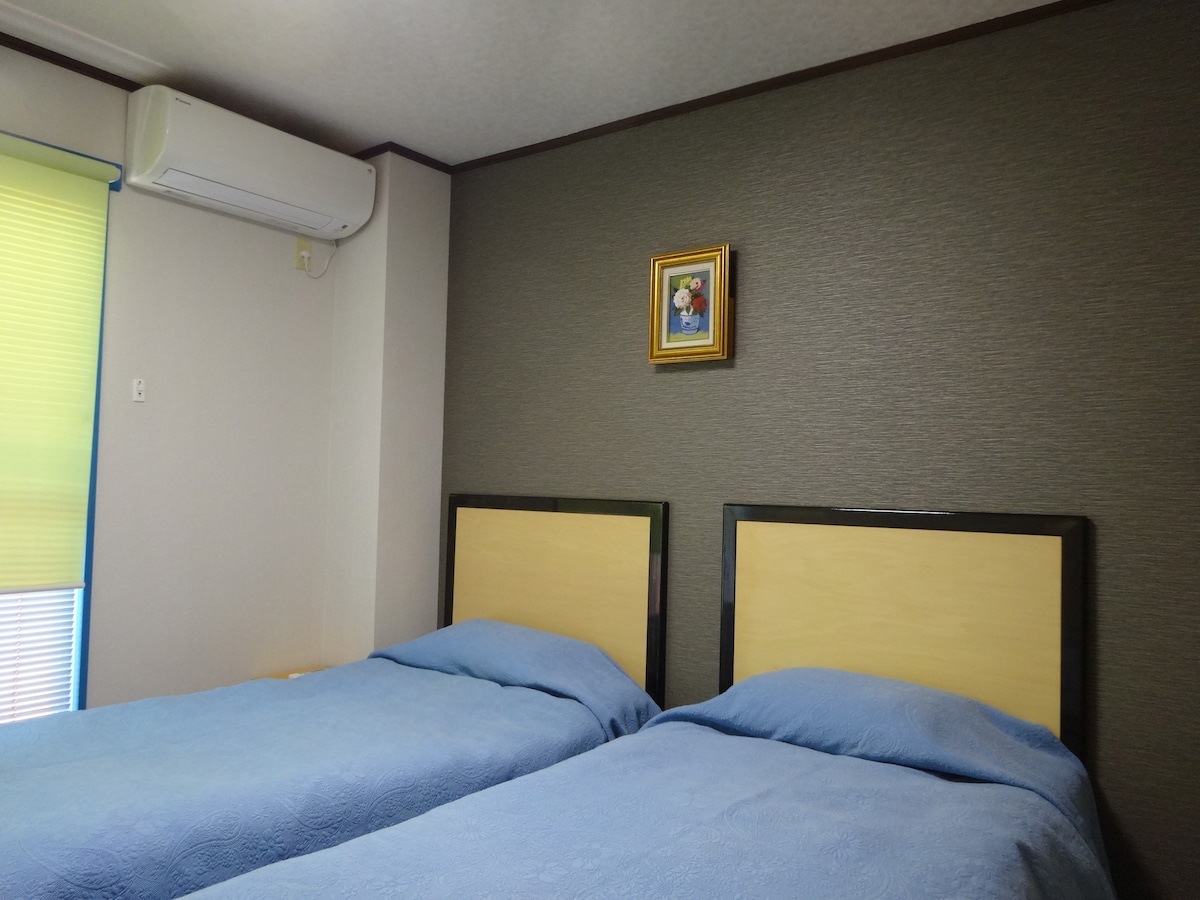 A room is shown with two spacious beds, each covered in light blue bedding. A framed artwork is mounted on the textured wall above. An air conditioning unit is positioned near a window adorned with light-filtering blinds.