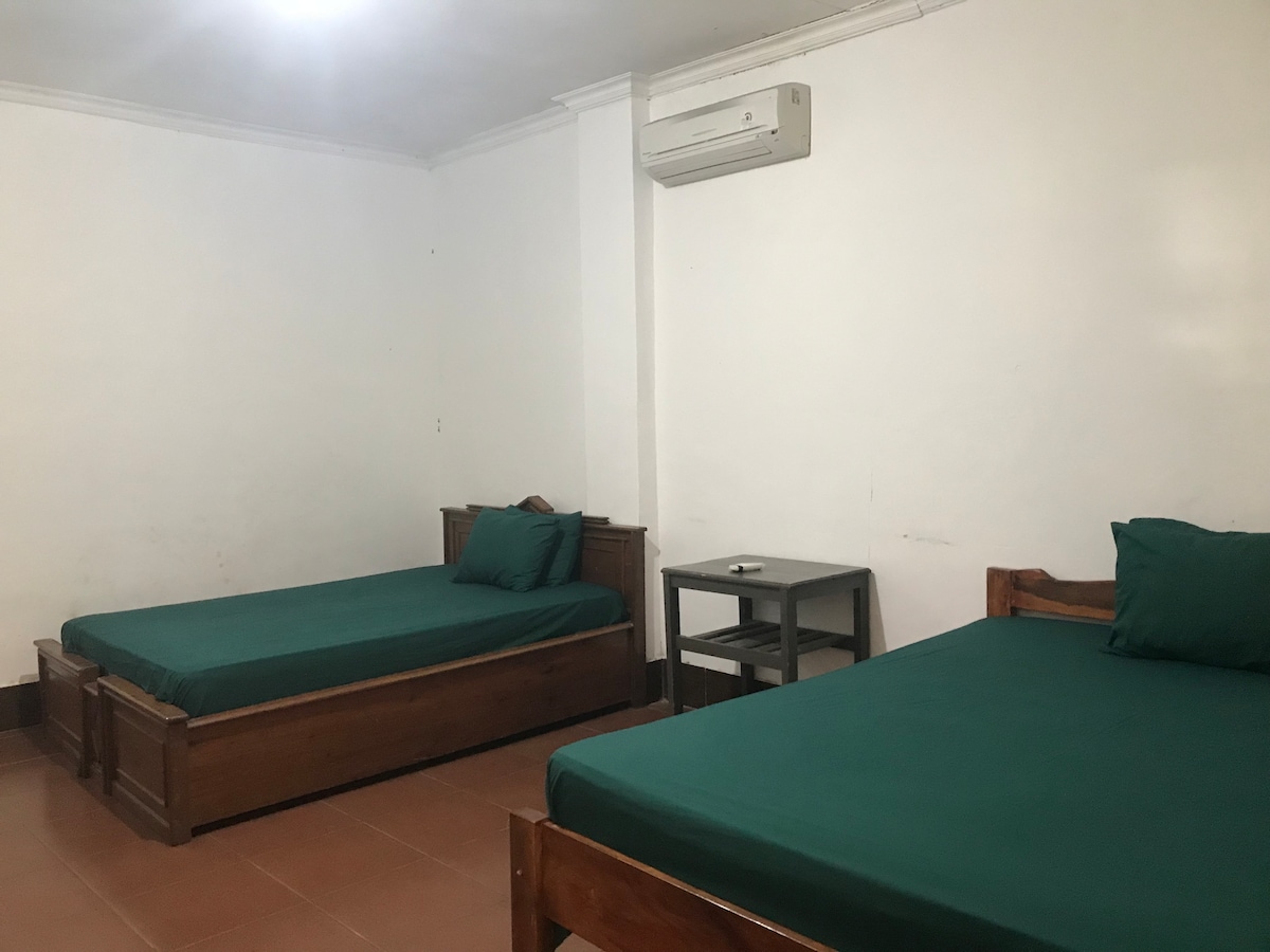 The room features two spacious wooden beds covered with green linens. A small table stands between the beds. A wall-mounted air conditioning unit is visible, along with plain white walls, creating a calm environment. Soft lighting illuminates the room.
