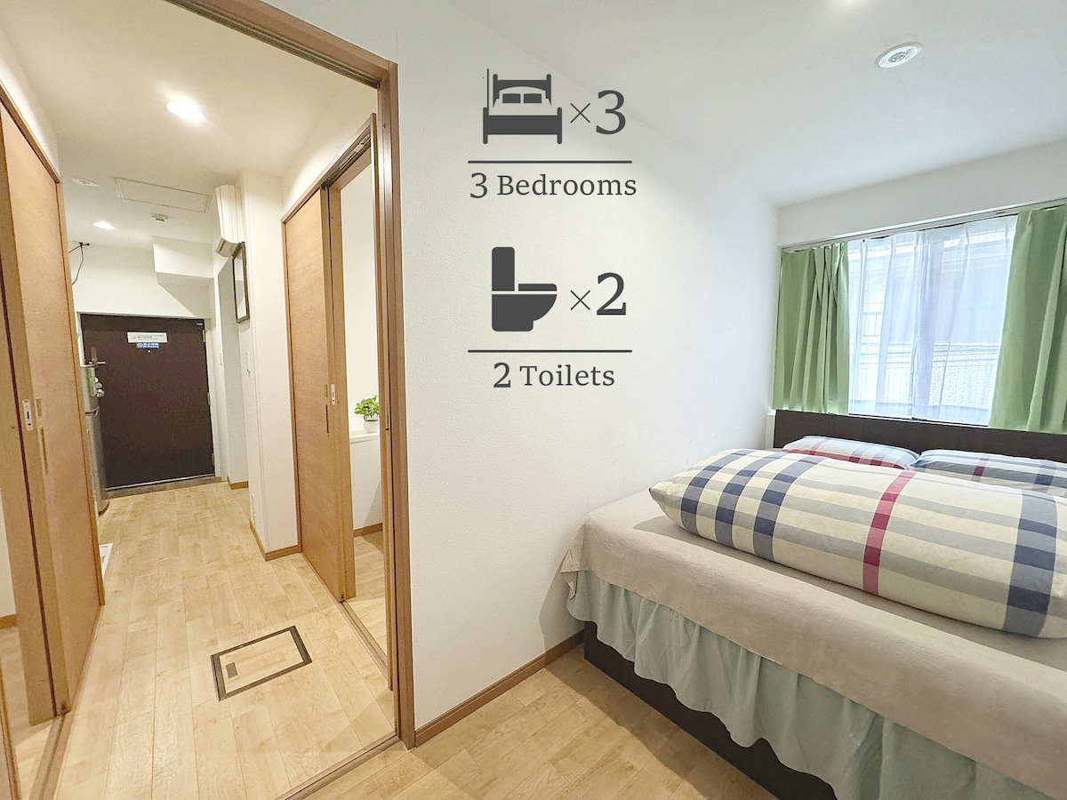 A bright room features a spacious bed with a plaid blanket. Two doors lead to adjacent areas, with a window draped in green curtains allowing natural light to fill the space. Icons indicate three bedrooms and two toilets for convenience.