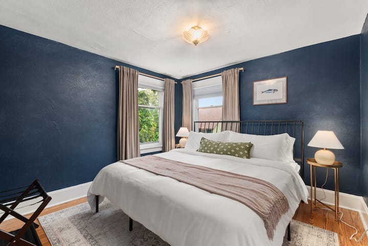 Მოიწვიეთ საძინებელი დიდი საწოლით, დამამშვიდებელი ლურჯი კედლებით და ბევრი ბუნებრივი განათებით ორი ფანჯრიდან.