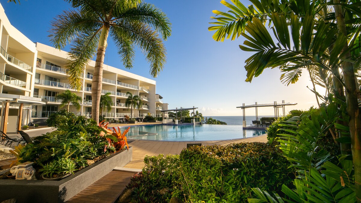 A spacious pool area is surrounded by lush tropical landscaping and palm trees. The modern building structure, featuring multiple balconies, is visible in the background, with the serene ocean horizon beyond. Sunlounges are arranged along the poolside for relaxation.