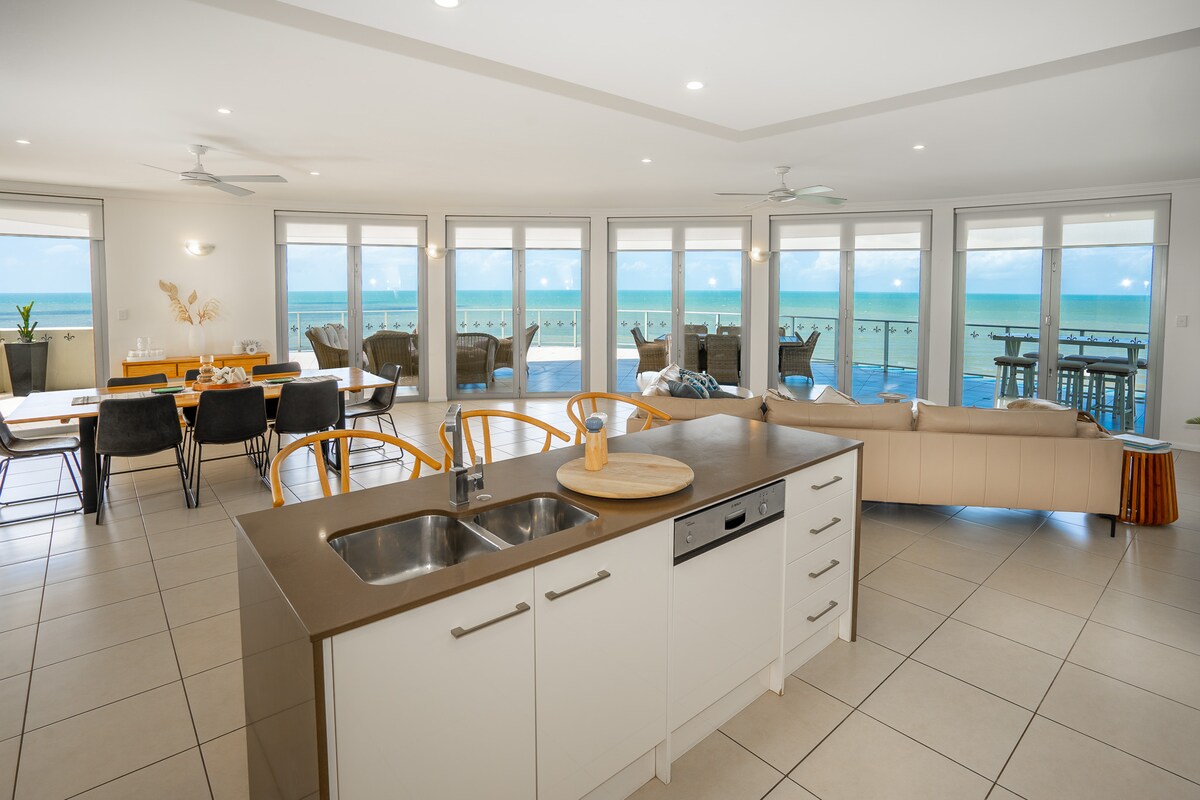 An open-plan living area showcases a modern kitchen with sleek cabinetry and stainless-steel appliances. Large windows frame panoramic ocean views, allowing natural light to fill the space. A dining table and comfortable seating area invite relaxation and enjoyment of the coastal scenery.