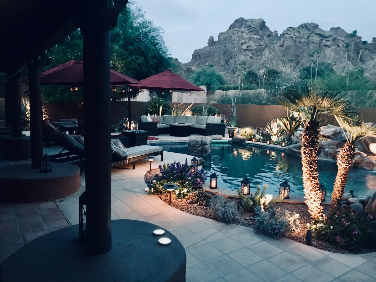 The serene backyard features a sparkling pool surrounded by lush landscaping, including palm trees and blooming flowers. Soft lighting from lanterns creates a cozy atmosphere, with comfortable seating areas under red umbrellas, all framed by the striking outline of the nearby mountains.