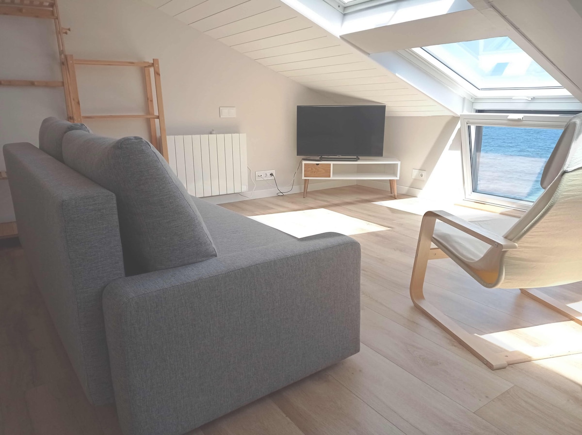 A comfortable seating area is featured with a light grey sofa positioned against a sloped wall. A flat-screen television stands on a white media console. Natural light floods the space through large skylights, illuminating the wooden floor and offering views of the ocean.