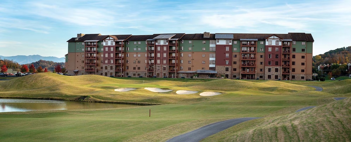 A multi-story resort building is visible, featuring a combination of earthy tones and balconies. The backdrop includes rolling hills and a clear sky. A golf course lies in the foreground with sand traps and a small pond, enhancing the landscape's natural appearance.