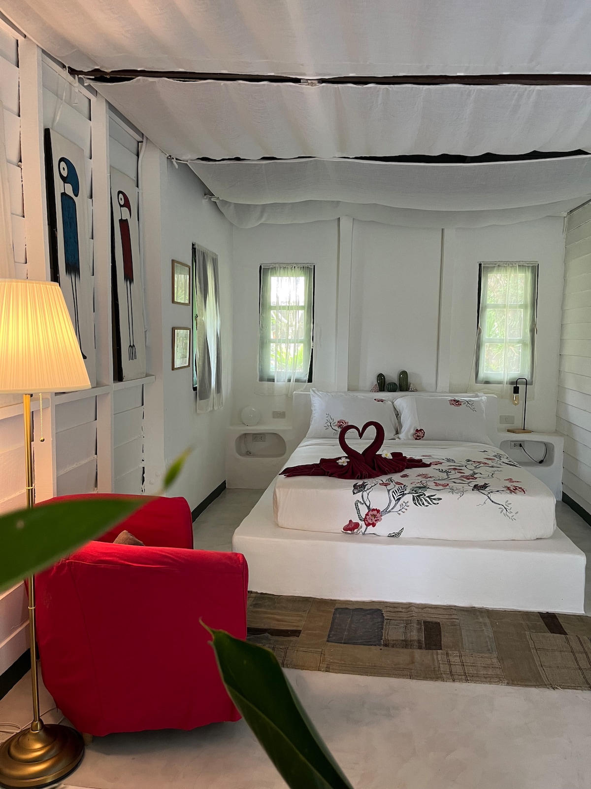 A bright sleeping area features a spacious bed adorned with floral accents and a decorative heart-shaped towel arrangement. Natural light filters through two windows beside the bed, creating a serene ambiance. A cozy red chair complements the minimalist decor, enhancing the inviting atmosphere.