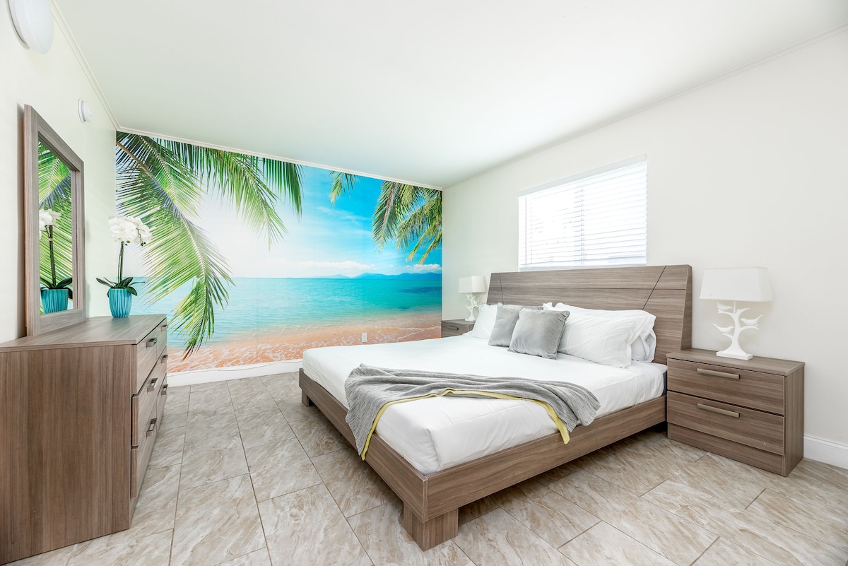 A spacious bedroom features a large bed with a soft blanket. A vibrant mural of a beach scene with palm trees adorns one wall. Two bedside tables with lamps are placed next to the bed, and a dresser is visible across from it.