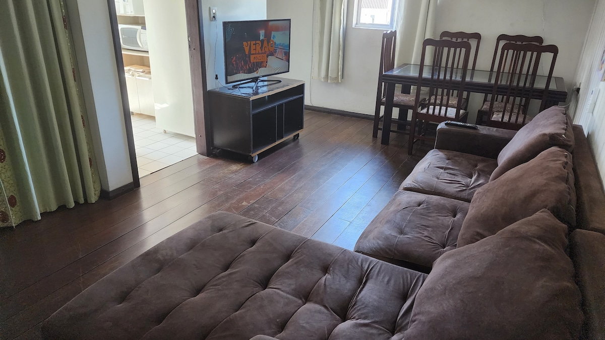 A spacious living area is furnished with a large sectional sofa and a dining table that accommodates several chairs. A television stands on a low black unit, while natural light enters through the nearby window, illuminating the wooden floor.