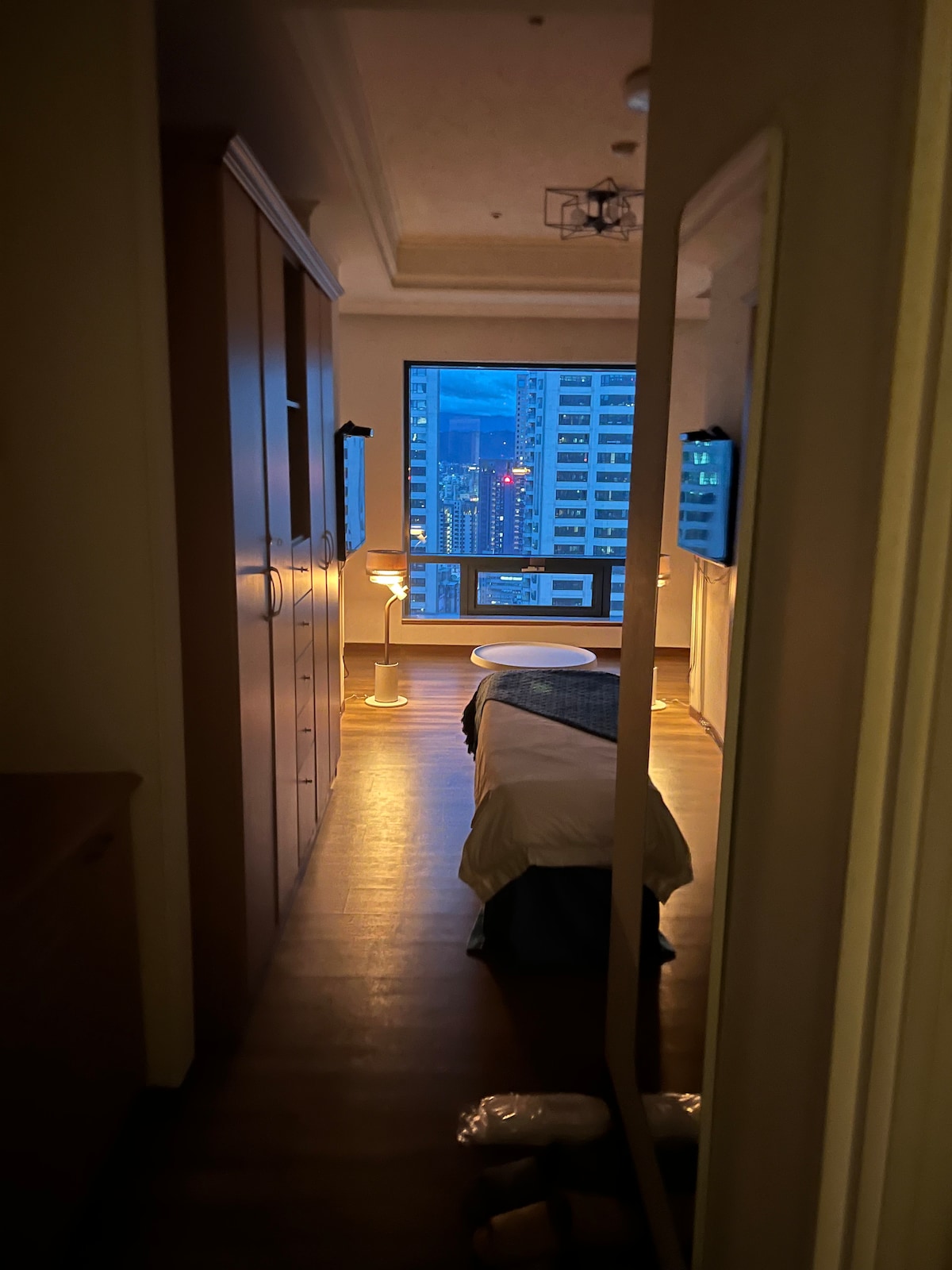 A narrow corridor leads to a well-lit bedroom featuring a spacious bed and bedside tables with lamps. A large window offers a view of the city skyline, where city lights begin to twinkle as evening falls.