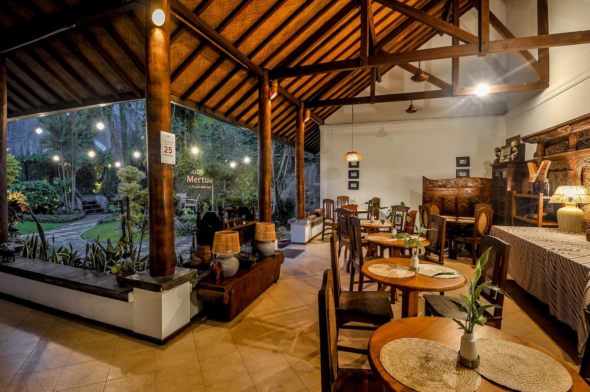 An intimate dining area is featured, showcasing wooden tables and chairs arranged around woven placemats. The space is illuminated by pendant lights, highlighting the traditional Javanese architecture and providing views of the lush garden outside through large glass windows.