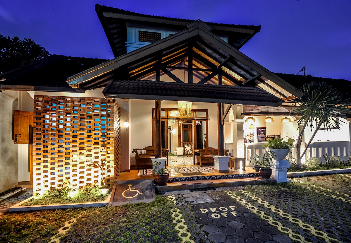 The exterior of the property features traditional Javanese architecture with a wooden door and large windows. A decorative brick wall provides visual interest, while potted plants add greenery. A designated 'Drop Off' area is marked on the paved pathway leading to the entrance.