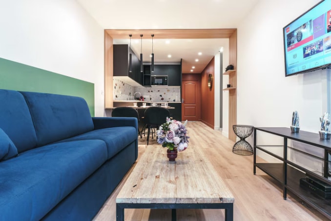 Dhomë ndenjjeje e rehatshme me divan krevat dhe televizor inteligjent, me pamje nga kuzhina e hapur 