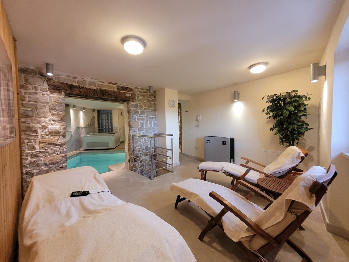 A wellness space is seen with stone walls and soft lighting, featuring a small swimming pool. Two loungers are positioned beside the pool, while a folding table adds functionality. The atmosphere is calm, complemented by a decorative plant in the corner.