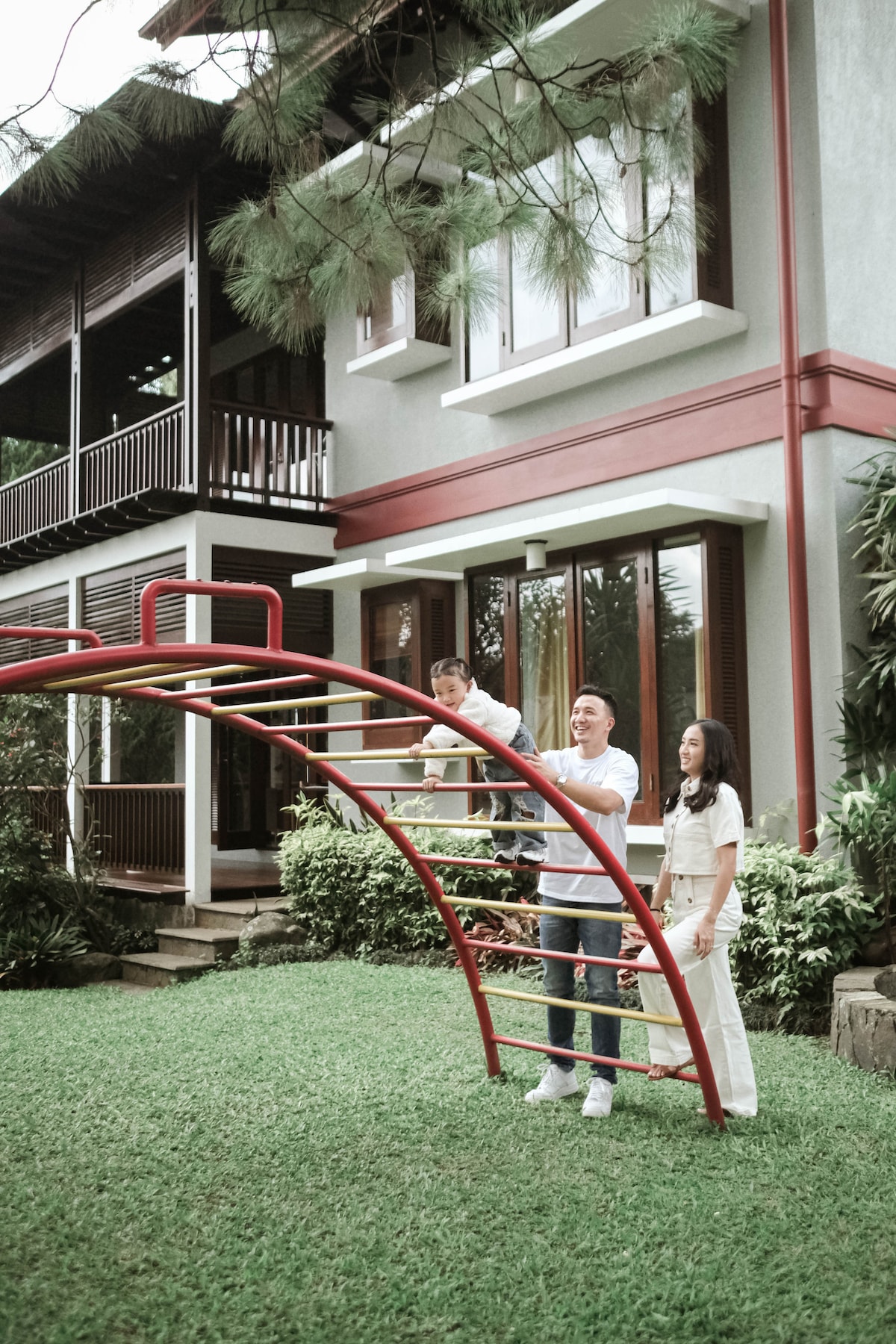 A red playground structure is positioned on lush green grass, surrounded by well-maintained plants. Two adults interact with a child nearby, while a two-story house with multiple windows and a balcony is visible in the background.