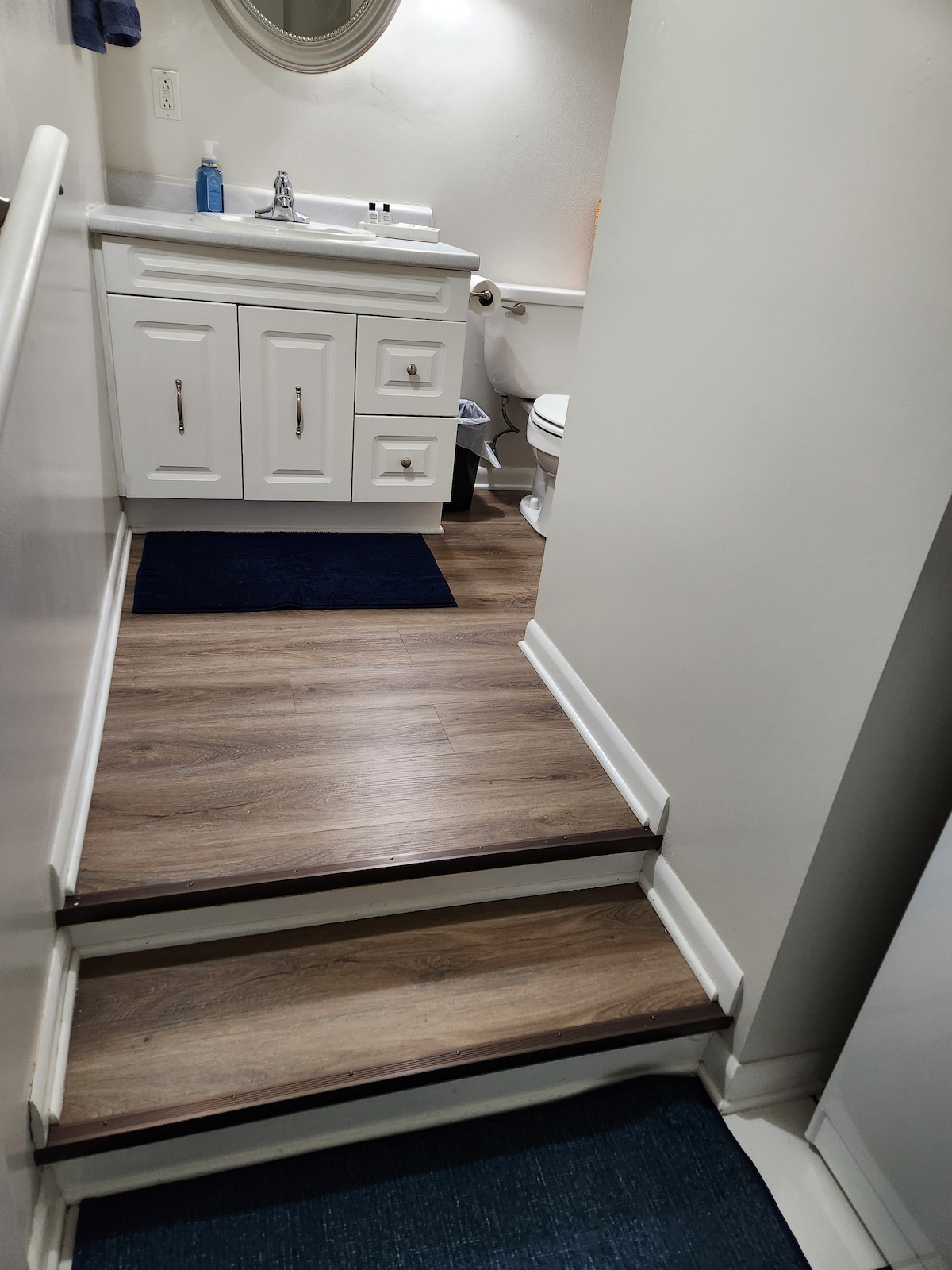 A staircase leads up to a bright bathroom featuring a white vanity with drawers and a round mirror. A blue mat is placed on the floor, complementing the warm wood tones of the stairs. A toilet is visible in the background, enhancing the functional design.