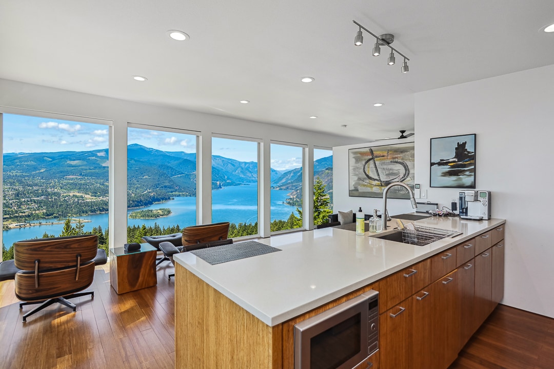 A modern kitchen area is showcased with sleek cabinetry and a large countertop. Expansive windows frame stunning views of the water and mountains. Comfortable seating is arranged nearby, allowing guests to enjoy the serene landscape from inside.