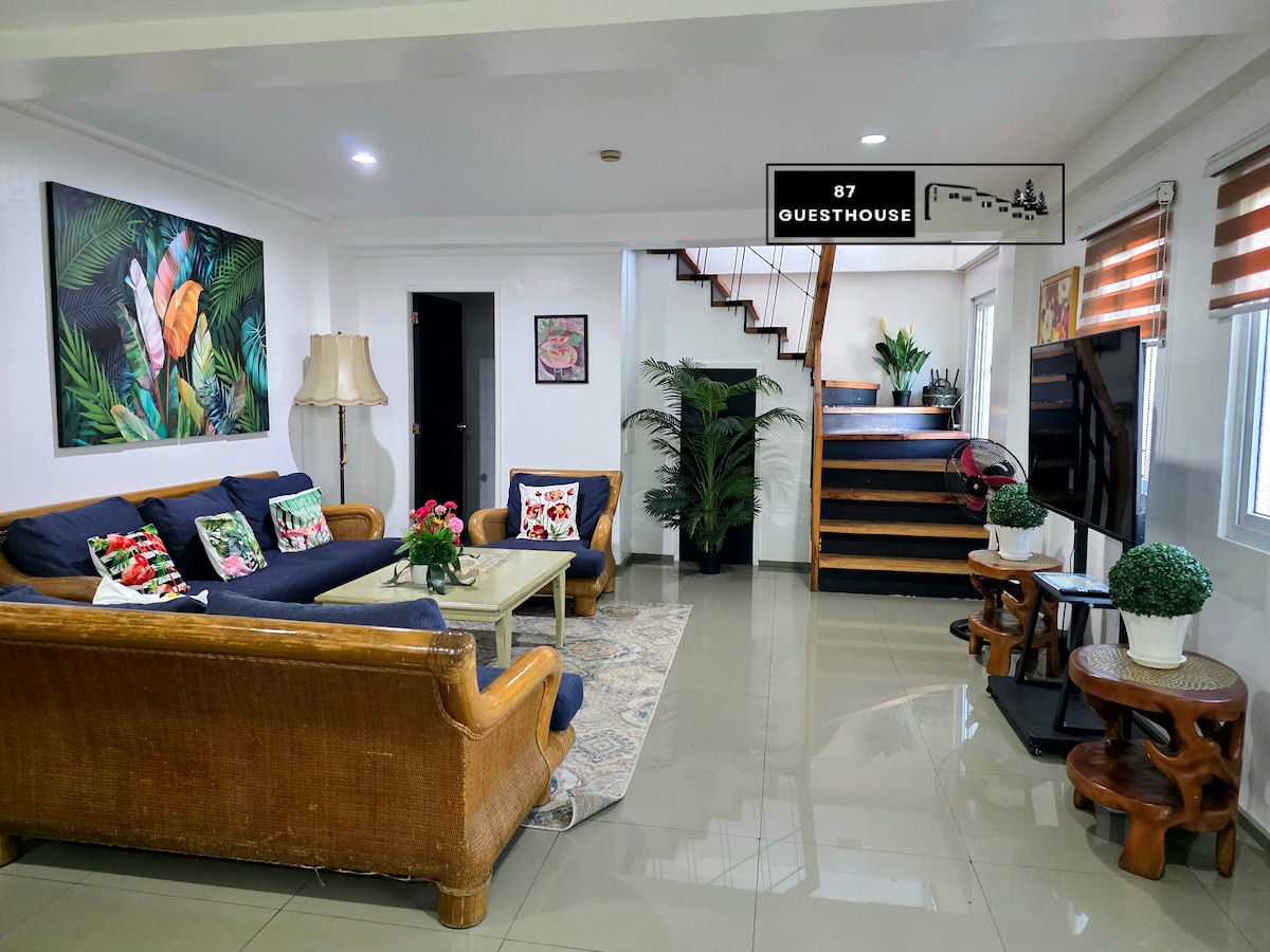 A cozy living area features a dark sofa adorned with colorful pillows and a large floor lamp. A woven rug lies beneath, and decorative plants add a touch of greenery. A wooden staircase ascends to the upper level, and wall art complements the bright ambience.