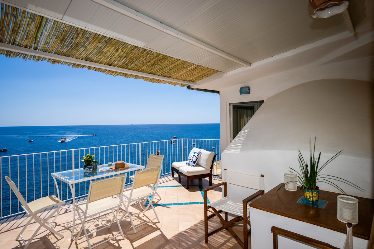 A private terrace overlooks the sea, featuring a glass dining table surrounded by four chairs. A comfortable sofa provides additional seating. A straw sunshade offers protection from sunlight, while the tranquil blue water is visible in the background, creating a serene atmosphere.