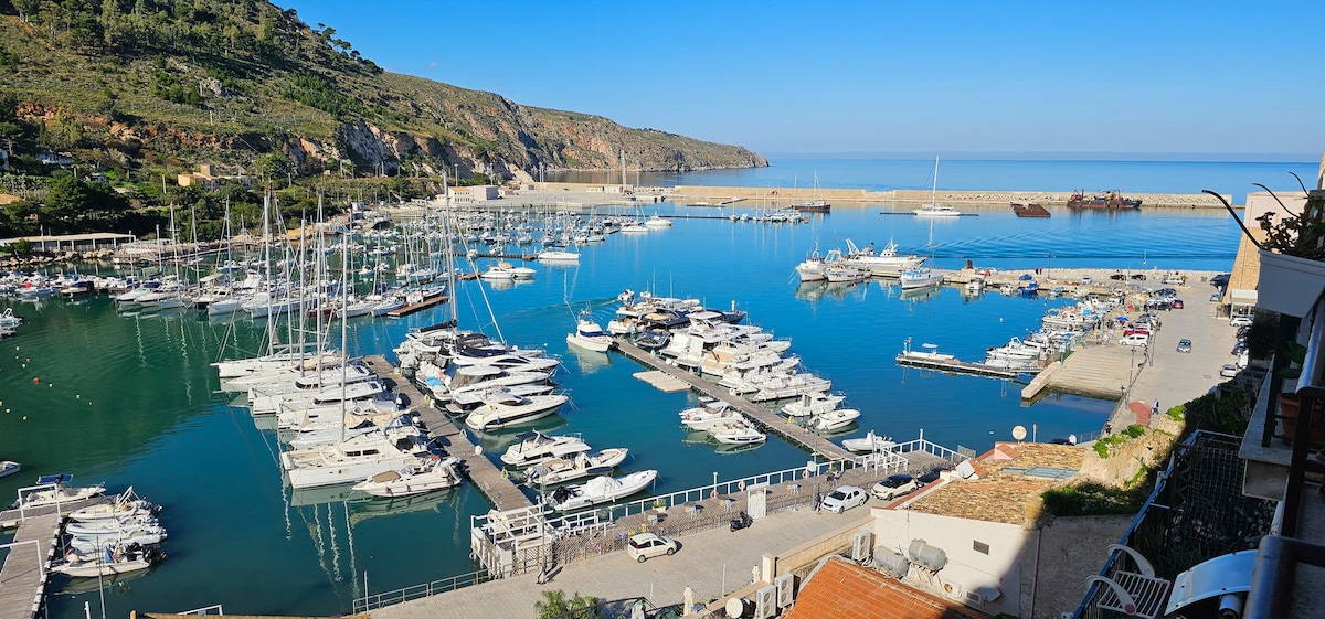 A panoramic view of a marina filled with numerous boats and yachts is shown, set against a serene blue sea. In the background, gentle hills and a clear sky create a tranquil atmosphere, while the harbor facilities are visible along the shoreline.