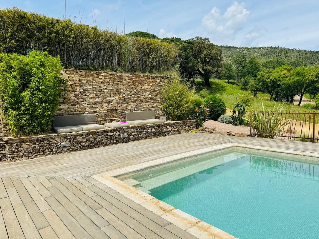 A refreshing salt-water pool is surrounded by natural landscaping and wooden decking. Comfortable seating is arranged on a stone terrace, providing a serene space to relax while enjoying the greenery and gentle slopes in the background.