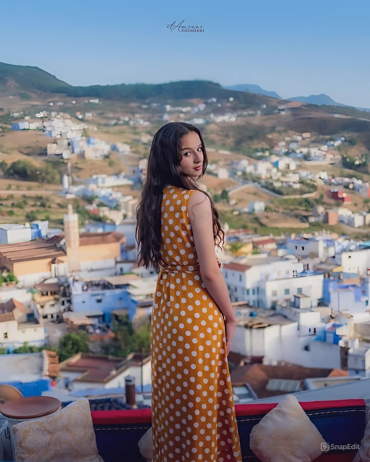 A figure stands on a terrace overlooking the colorful buildings of Chefchaouen’s Medina. The view captures layers of white and blue structures bordered by mountains and a bright sky. Soft hues of the landscape create a blend of natural beauty.