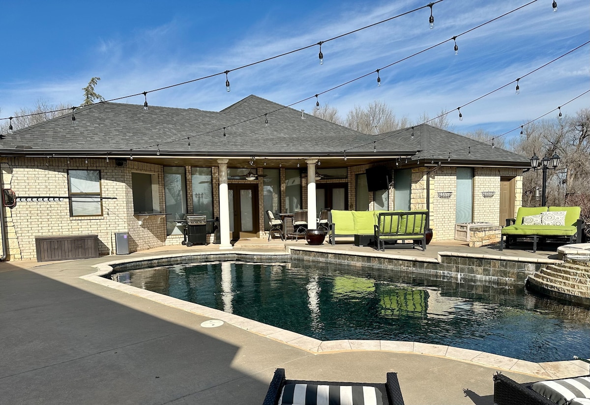 A spacious outdoor area showcases a large pool surrounded by a concrete patio. Comfortable seating arrangements are visible, including loungers and sofas. The home features multiple entry points, and string lights are draped overhead, enhancing the ambiance.