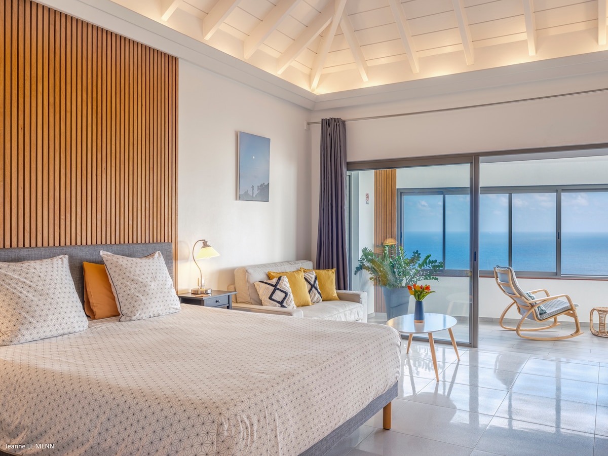 A spacious bedroom features a queen-size bed adorned with patterned linens and two pillows. A seating area includes a sofa and a chair, with natural light flooding in through large glass doors that offer views of the sea.