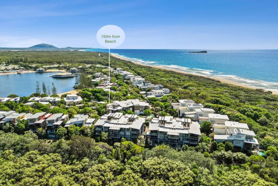 An aerial view showcases the resort's layout, with proximity to the beach clearly indicated. Lush greenery surrounds the buildings, and the coastline stretches out in the distance. The beach, lagoon, and surrounding nature integrate harmoniously into the landscape.