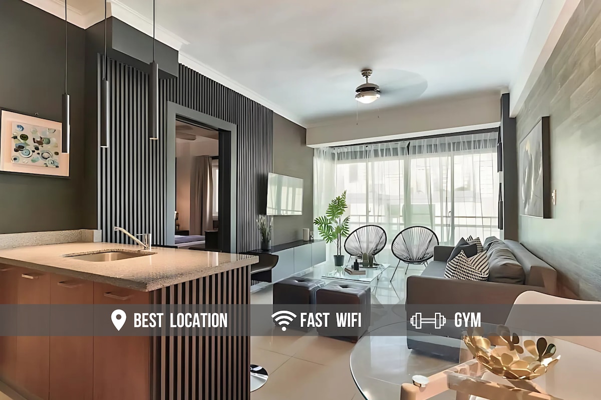 An open concept living area is visible, showcasing a modern kitchen with a breakfast bar and sleek cabinetry. Comfortable seating arrangements include a sofa and lounge chairs, while large windows allow natural light to fill the space, enhancing the airy feel. Decorative artwork adorns the walls.