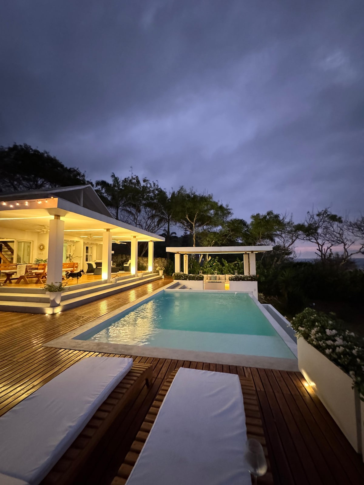 A serene outdoor space features a sparkling pool surrounded by a wooden deck. Comfortable lounge chairs line the edge of the pool, while the covered patio offers a dining area. Soft lighting enhances the tranquil atmosphere against a twilight sky.