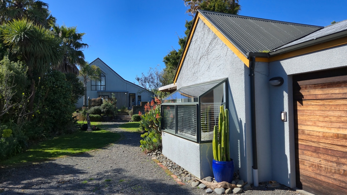 A landscaped pathway leads through a garden adorned with native plants, creating a serene entryway to Cheesemakers Retreat. The self-contained studio is framed by vibrant foliage, with a blue planter showcasing a tall succulent beside the entrance. Clear skies and sunlight enhance the inviting atmosphere.