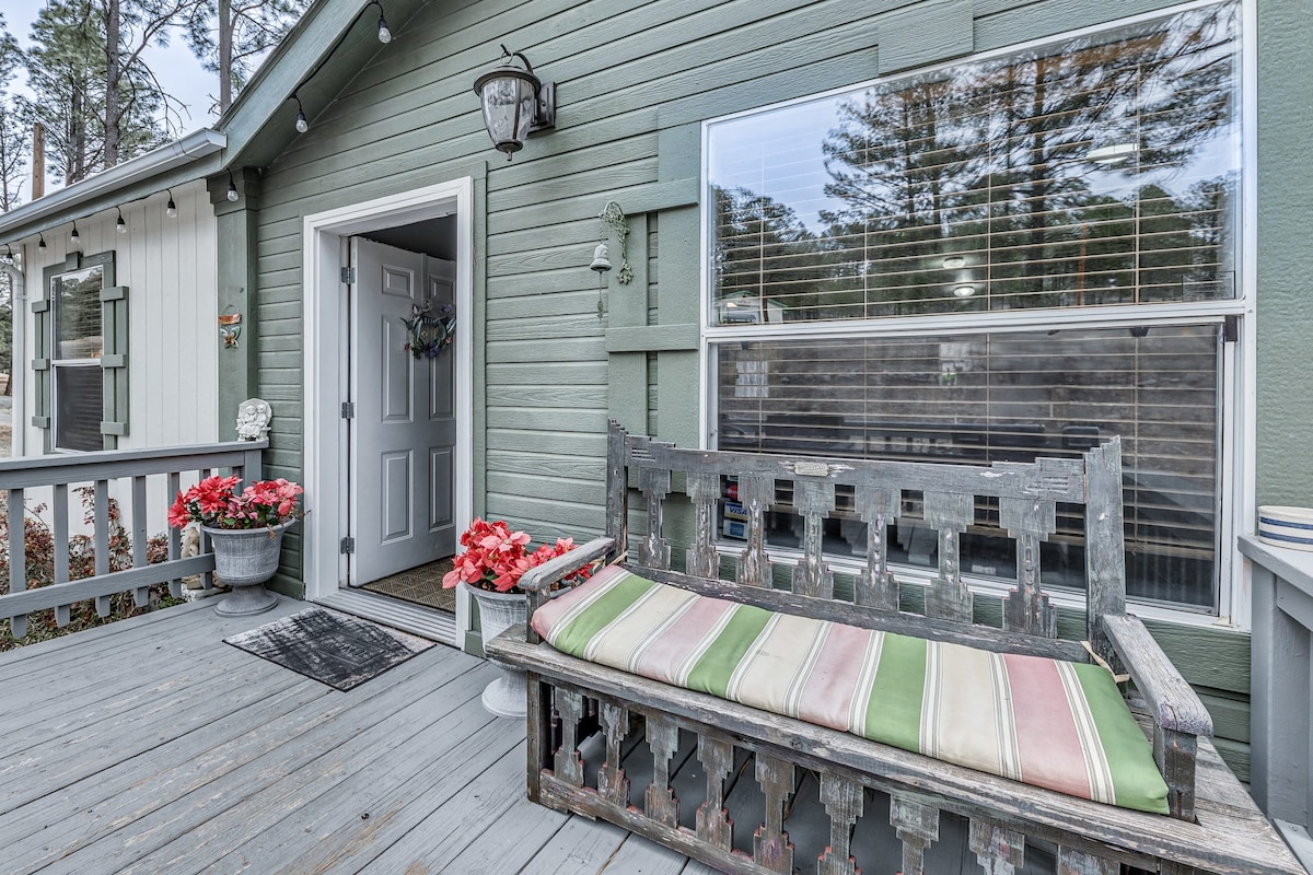 A welcoming front porch features a decorative wooden bench with a multi-striped cushion, surrounded by planters filled with vibrant flowers. A front door is visible, providing access to the interior, with large windows allowing natural light to illuminate the space.