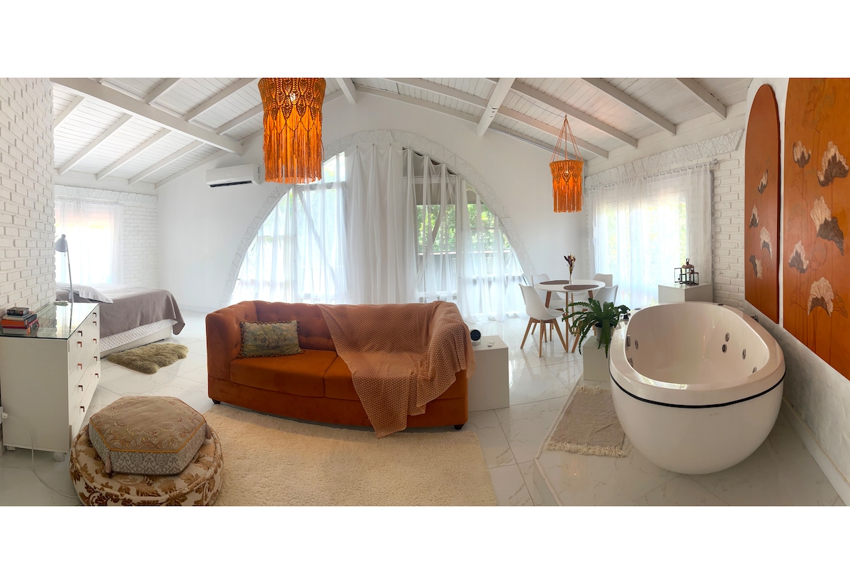 A spacious interior is highlighted by natural light filtering through large windows. An inviting sofa with a soft throw is positioned alongside a modern freestanding tub, while warm lighting fixtures add a gentle glow to the room's contemporary design. The area is adorned with subtle decorative elements, enhancing the overall atmosphere.