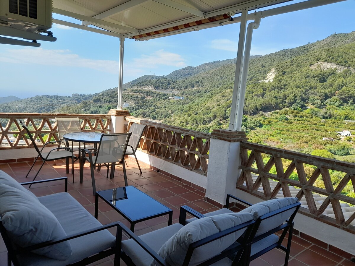 A spacious terrace is furnished with a comfortable sofa set and a small dining table, offering a serene outdoor space. Scenic mountain views are visible in the background, while a canopy provides shelter from the sun. The floor features terracotta tiles adding warmth to the area.