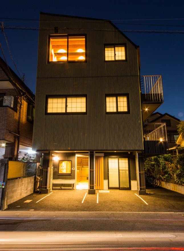 The exterior of a three-story private building is illuminated at night, showcasing its unique design with a combination of dark-colored panels and large window sections. Parking spaces are visible in front, while the surroundings reflect a quiet residential area with minimal traffic.