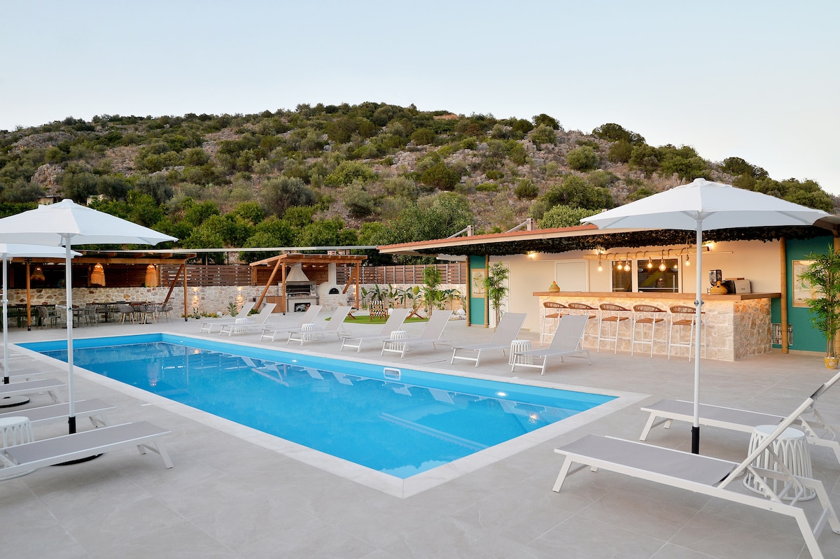 A bright swimming pool is surrounded by sunbeds and umbrellas, with a stone bar and seating area visible in the background. Lush hills provide a natural backdrop, creating a relaxing atmosphere for guests to enjoy.