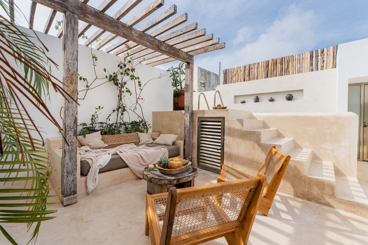 An open-air terrace features a comfortable seating area adorned with greenery, complemented by wooden furniture. A cozy sofa is draped with a soft blanket, while a rustic table holds fruit. Steps lead to a higher level, enhancing the rooftop experience.