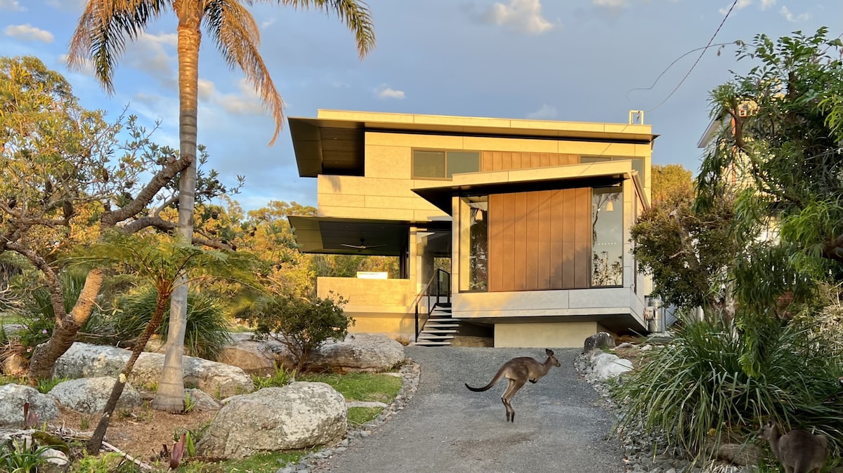 The exterior of the house is showcased with modern architectural lines and large windows. A pathway leads up to the entrance, surrounded by native plants and beach stones. A kangaroo can be seen gracefully crossing the path, highlighting the natural surroundings.