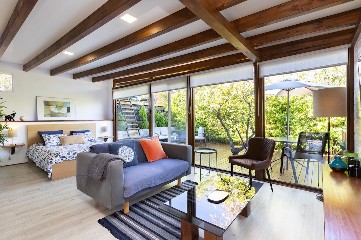 A modern living space is illuminated by natural light, highlighting a comfortable sofa with an orange and gray accent. An inviting queen-size bed is seen in the background, while large glass doors open to a terrace surrounded by greenery.