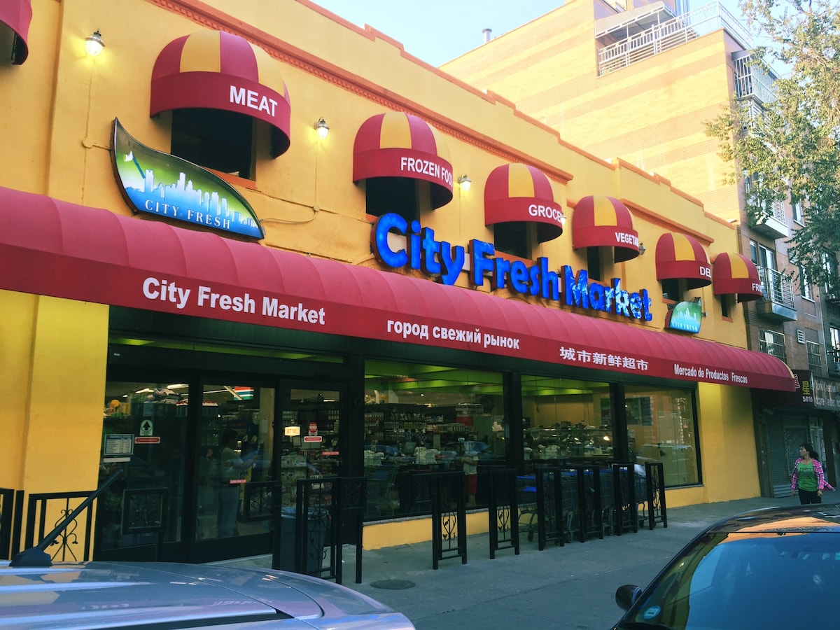 A vibrant storefront of City Fresh Market is displayed, featuring a colorful yellow exterior and large red awnings. The signage exhibits the store's name in English and multiple languages. Various sections for meat, groceries, and frozen items are clearly identified, inviting shoppers.